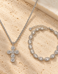 Silver cross pendant necklace and matching beaded bracelet with clear stones, displayed on a beige stone surface.