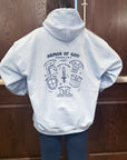 Person wearing a light blue hoodie with 'Armor of God' design on the back, standing against a wooden door.