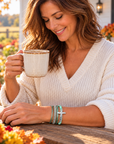 A woman in a cozy white knit sweater sits at a wooden table outdoors, holding a ceramic mug. She is wearing a stack of silver-toned bracelets, including a cross charm bracelet and a green beaded bracelet, on her wrist.