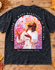 A black acid-wash graphic t-shirt laid flat on a rustic wooden background decorated with autumn leaves, a small pumpkin, and dried flowers. The back of the shirt features a large colorful graphic of Jesus in a white robe surrounded by pink and red hearts, with the text 'Everything is made beautiful in His time' and 'Ecclesiastes 3:11'.