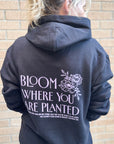Person wearing a black hoodie with 'Bloom Where You Are Planted' text and floral design on the back, standing against a brick wall.