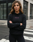 Woman wearing a black hoodie with text, standing on a street with a building in the background