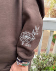 Brown sweatshirt with floral design on sleeve, held by a person wearing colorful bracelets.
