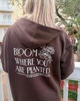 Person wearing a brown hoodie with 'Bloom Where You Are Planted' text and floral design on the back, standing outdoors.