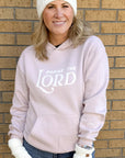 Person wearing a light pink hoodie with 'Praise the Lord' text, white beanie, and gloves against a brick wall.