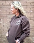 Woman wearing a brown hoodie with text, standing against a brick wall.