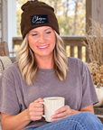 A smiling woman sits on a porch wearing a brown knit beanie with a black patch that reads 'Chosen 1 Peter 2:9' and holding a speckled cream mug. She wears a faded mauve-gray t-shirt and blue jeans, with a rustic wooden chair and fall decorations in the background.