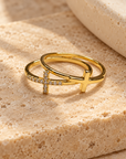 Two gold rings displayed on a textured beige stone surface. One ring features a polished cross with a row of small clear stones, and the other is a smooth open band.