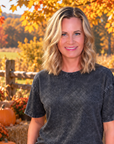 A woman stands outdoors in a fall yard wearing a faded charcoal gray short-sleeve t-shirt and blue jeans. Pumpkins, mums, a wooden fence, and autumn trees are visible in the background.