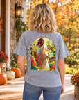 A woman stands on a porch wearing a heather gray graphic T-shirt from the back, featuring a colorful print of Jesus in a garden with the text 'he has made everything beautiful in its time