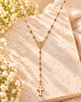A delicate gold Y-necklace with a heart-shaped medallion and cross pendant, adorned with small multicolored beads, displayed on a textured beige stone slab with white flowers and cream fabric.