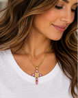 A gold cross pendant necklace with pink gemstones and a small central religious medallion, shown on a white background and worn by a woman in a white top.