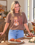 A woman wearing a brown graphic t-shirt with a fall-themed teddy bear design and the text 'GIVE THANKS TO THE LORD' standing in a cozy, decorated dining room.