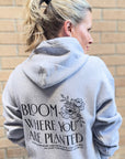 Person wearing a gray hoodie with 'Bloom Where You Are Planted' text and floral design on the back, standing against a brick wall.