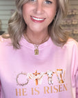 Woman wearing a pink sweatshirt with 'He is Risen' text and religious symbols.