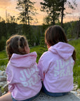 Two children wearing pink hoodies with 'Just Pray' text, sitting on a rock in a natural setting.
