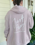 Person wearing a light pink hoodie with 'Just Pray' text on the back, standing outside.