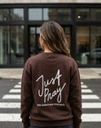 Person wearing a brown sweatshirt with 'Just Pray' text on the back, standing on a crosswalk.