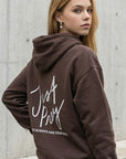 Person wearing a brown hoodie with 'Just Pray' text on the back, standing against a concrete wall.