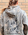 A woman viewed from behind wearing a gray and white distressed camouflage hoodie with 'Just Pray' and 'GOD ALWAYS HAS YOUR BACK.' printed in black on the back, standing against a tan brick wall.