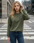 Woman wearing an olive green sweatshirt with text and blue jeans on a city street.