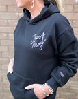 Person wearing a black hoodie with 'Just Pray' text, standing against a brick wall.