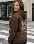 Woman wearing a brown hoodie with text on the back, standing on a city street.