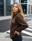 Woman wearing a brown hoodie with text, standing on a city street.