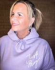 Woman wearing a lavender hoodie with 'Just Pray' text against a brick wall.