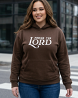 Woman wearing a brown hoodie with 'Praise the Lord' text, standing on a street.