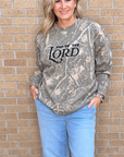 A woman wearing a long-sleeve camouflage sweatshirt with 'PRAISE THE LORD' printed in black across the chest, paired with light blue jeans, standing against a tan brick wall.