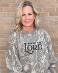A woman wearing a long-sleeve crewneck sweatshirt with a brown and beige camouflage pattern and 'PRAISE THE LORD' printed in black across the chest, paired with light blue jeans, standing against a tan brick wall.