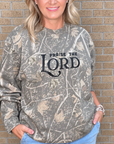 A woman wearing a camouflage-patterned long-sleeve sweatshirt with 'PRaise THE LORD' printed in black text on the front, paired with light blue jeans, standing against a tan brick wall.
