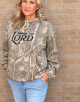 A woman wearing a camouflage hoodie with 'PRaise THE LORD' printed in black across the chest, paired with light blue jeans, standing against a tan brick wall.