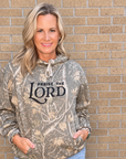 A woman wearing a camouflage hoodie with 'PRAISE THE LORD' printed on the front, paired with light blue jeans, standing against a tan brick wall.