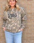 A woman wearing a camouflage hoodie with 'PRaise THE LORD' printed in black across the chest, paired with light blue jeans, standing against a tan brick wall.