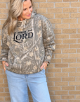 A woman wearing a camouflage hoodie with 'PRaise THE LORD' printed in black on the chest, paired with light blue jeans, standing against a tan brick wall.