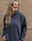 A woman with long blonde hair smiling and looking to the side, wearing a dark charcoal gray oversized hoodie and blue jeans, standing in front of a brown brick wall.
