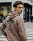 Man wearing a beige hoodie with a motivational message on the back, standing on a city street.