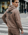 Man wearing a beige hoodie with a motivational message on the back, standing on a city street.