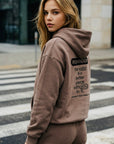 Woman wearing a brown hoodie with text on the back, standing on a crosswalk.