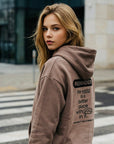 Woman wearing a brown hoodie with text on the back, standing on a crosswalk.