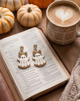 A pair of white ghost-shaped earrings with gold glittery bows and gold lettering, displayed on an open book in a cozy autumn-themed setting with pumpkins, a coffee cup, and a knit blanket.