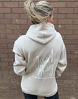 Person wearing a beige hoodie with 'Just Pray' text on the back against a brick wall.