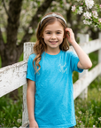 Young girl in a blue shirt standing in front of a white picket fence with cherry blossom trees in the background.