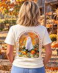 A woman stands in a pumpkin patch wearing a cream-colored graphic t-shirt. The back of the shirt features a large graphic of a white ghost surrounded by orange pumpkins and marigolds, with the text 'AIN'T NO GHOST LIKE THE HOLY GHOST' arched above the design.