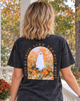 A woman stands on a white porch wearing a faded black graphic T-shirt from the back, showing a large design of a ghost in a field with autumn leaves and the text 'ain't NO GHOST LIKE THE holy GHOST ' She is also wearing blue jeans, with pumpkins and orange flowers on the porch beside her.