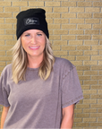 A woman with blonde hair wearing a black beanie with a white embroidered logo and a faded gray t-shirt, standing in front of a tan brick wall.