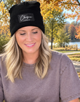 A woman sitting at a wooden table outdoors in a park, wearing a black beanie with a white 'Chosen' patch and Trees with autumn foliage and a lake are visible in the background.