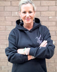 Woman wearing a black hoodie with text, standing against a brick wall.
